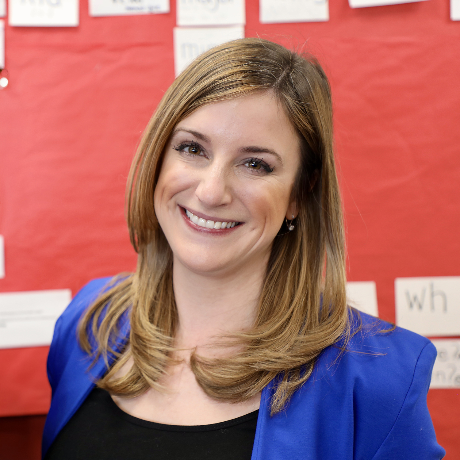 Portrait of Principal Kelly Espinosa from Fanny Jackson Coppin School, smiling and wearing a blue blazer in front of a red classroom bulletin board with word cards.