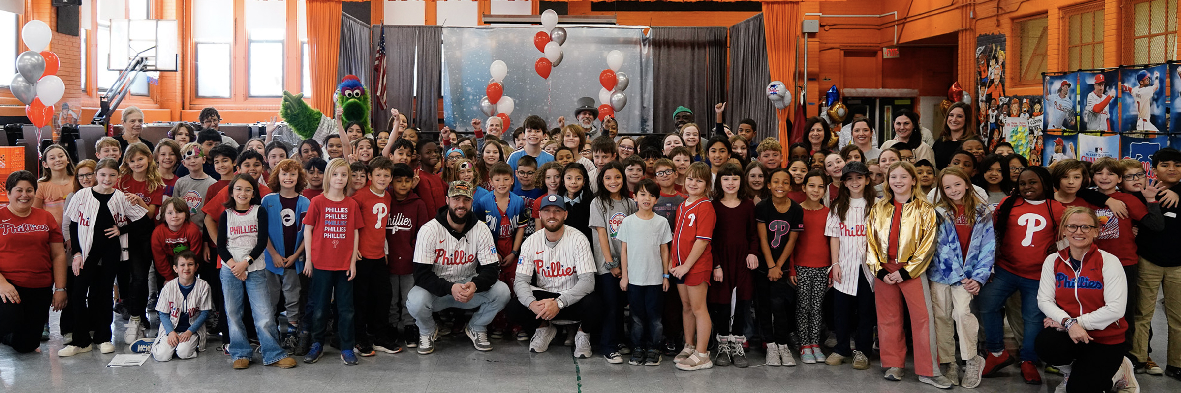 Large group of Fanny Jackson Coppin School students and staff gathered in the gym with Philadelphia Phillies players and the Phillie Phanatic mascot. Many participants wear Phillies shirts and jerseys, smiling and posing for the camera with balloons and posters in the background.